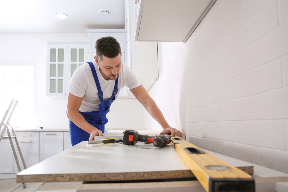 A man in overalls is busy at a kitchen countertop installation, concentrating on his work with various tools and items nearby.